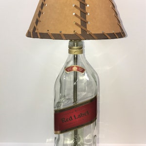 Puede incluir: Una l&aacute;mpara de mesa hecha de una botella de vidrio transparente con una pantalla de cuero marr&oacute;n. La botella tiene una etiqueta roja que dice "Johnnie Walker Red Label" y una etiqueta dorada que dice "1,75 Litros". La pantalla tiene costuras de cuero marr&oacute;n.