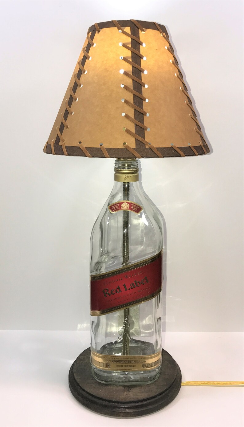 Puede incluir: Una l&aacute;mpara de mesa hecha de una botella de vidrio transparente con una pantalla de cuero marr&oacute;n. La botella tiene una etiqueta roja que dice "Johnnie Walker Red Label" y una etiqueta dorada que dice "1,75 Litros". La pantalla tiene costuras de cuero marr&oacute;n.