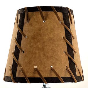 May include: A lampshade with a brown paper-like material and dark brown trim. The shade is stitched with tan-colored leather-like strips. The shade has a tapered shape and is designed to fit on a lamp base. The overall design is rustic and vintage.