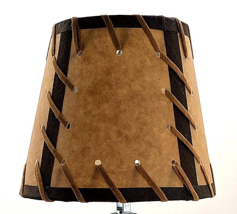 May include: A lampshade with a brown paper-like material and dark brown trim. The shade is stitched with tan-colored leather-like strips. The shade has a tapered shape and is designed to fit on a lamp base. The overall design is rustic and vintage.