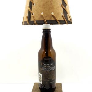 May include: A unique lamp crafted from a Guinness Extra Stout beer bottle. The lampshade is tan with dark brown trim and stitching. The lamp sits on a wooden base and has a brown power cord.