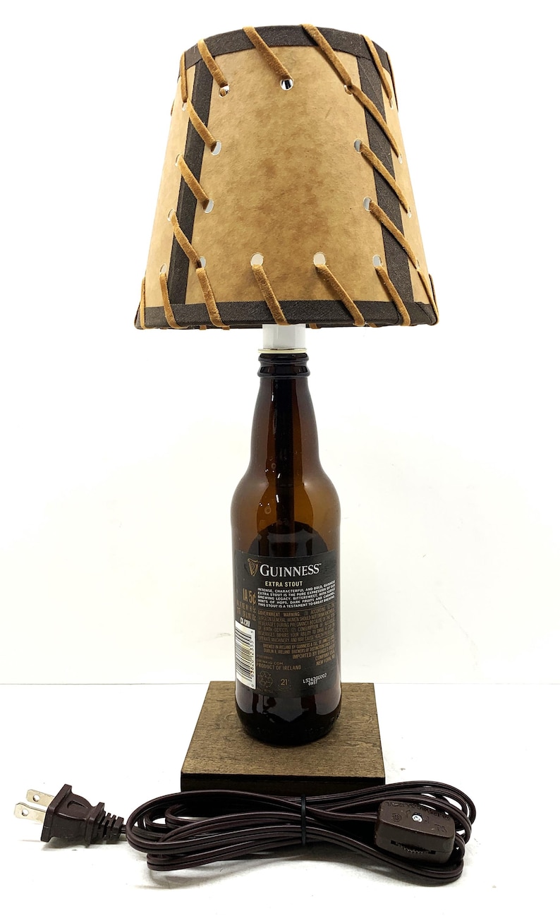 May include: A unique lamp crafted from a Guinness Extra Stout beer bottle. The lampshade is tan with dark brown trim and stitching. The lamp sits on a wooden base and has a brown power cord.