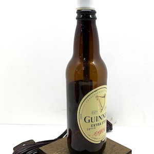 May include: A repurposed Guinness Extra Stout beer bottle lamp. The brown glass bottle has a white lamp shade and sits on a small, square wooden base. A brown electrical cord extends from the base.