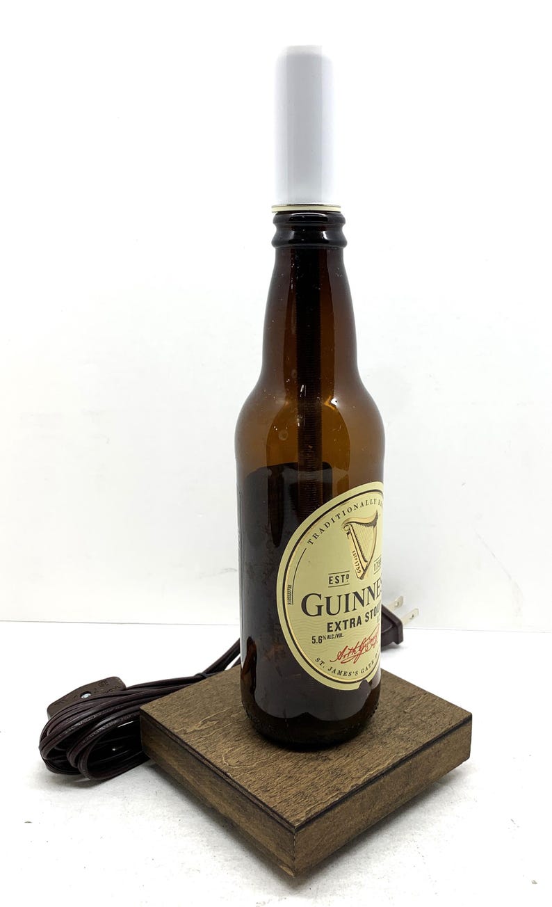 May include: A repurposed Guinness Extra Stout beer bottle lamp. The brown glass bottle has a white lamp shade and sits on a small, square wooden base. A brown electrical cord extends from the base.