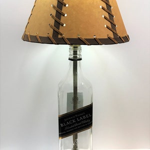 May include: A table lamp made from a repurposed Johnnie Walker Black Label Scotch whisky bottle. The lamp has a brown leather lampshade with a criss-cross pattern and a wooden base.