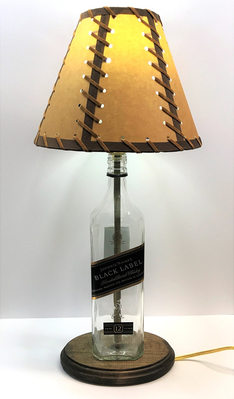 May include: A table lamp made from a repurposed Johnnie Walker Black Label Scotch whisky bottle. The lamp has a brown leather lampshade with a criss-cross pattern and a wooden base.