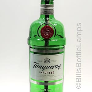 May include: A green glass bottle lamp with a gold metal lampshade. The bottle is labeled "Tanqueray Imported London Dry Gin" and has a red seal on the front. The lamp is sitting on a wooden base.