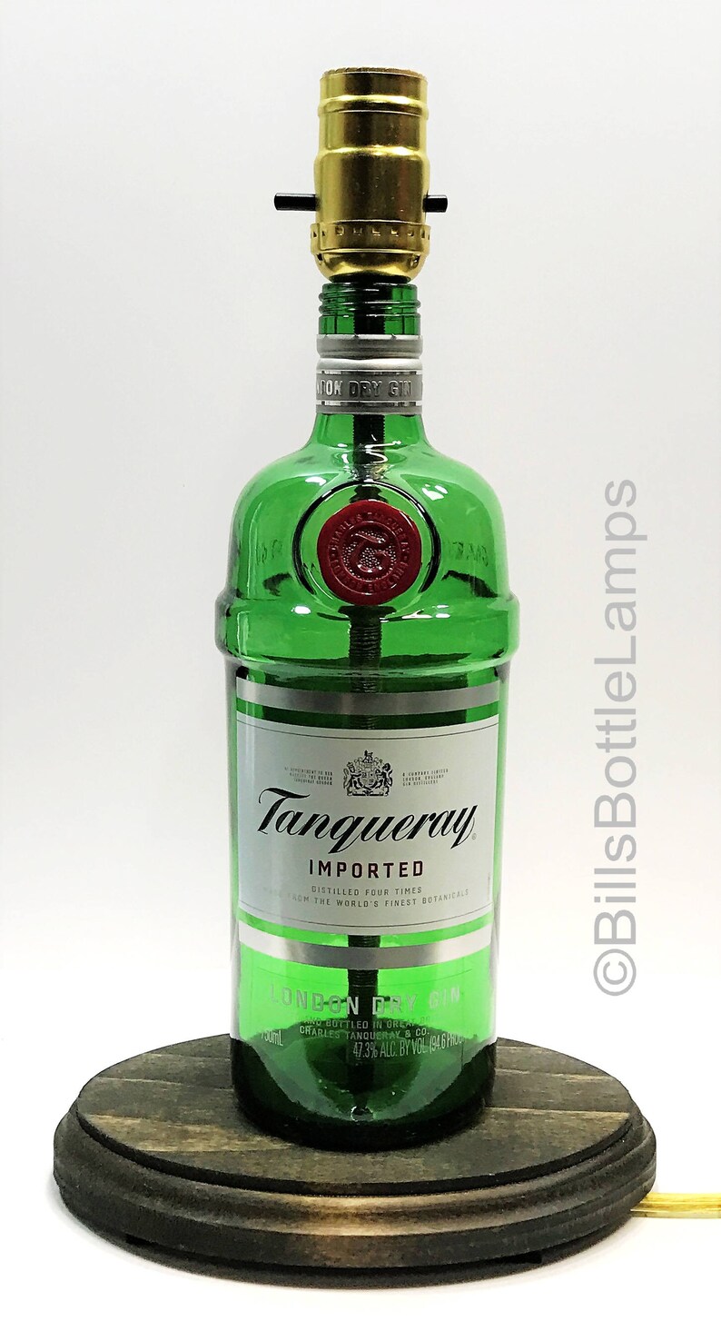 May include: A green glass bottle lamp with a gold metal lampshade. The bottle is labeled "Tanqueray Imported London Dry Gin" and has a red seal on the front. The lamp is sitting on a wooden base.