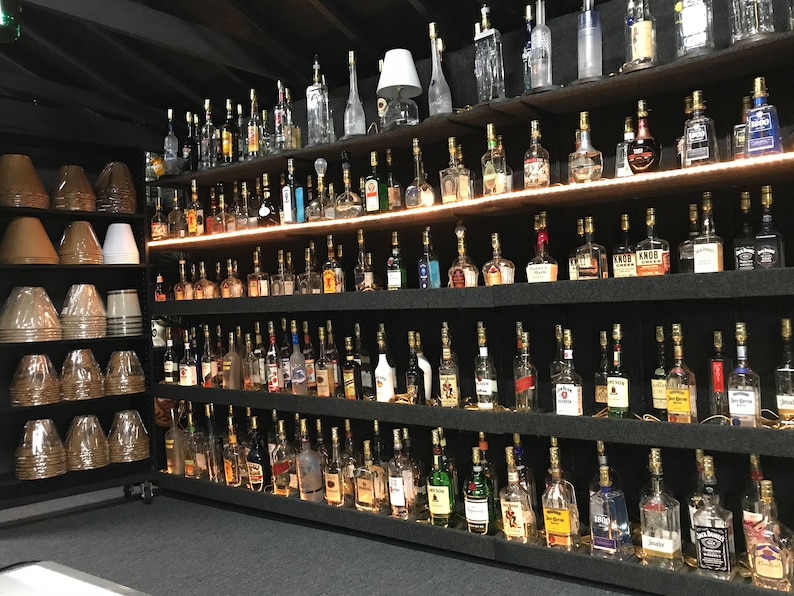 May include: A wall of shelves filled with glass bottles, each with a light bulb inside, creating a unique lighting display. The bottles are arranged in rows and columns, with a variety of shapes and sizes. The shelves are black and the bottles are clear glass.