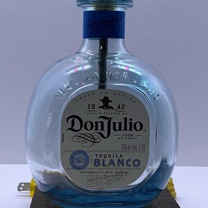 May include: A clear glass bottle lamp with a gold metal top. The bottle is labeled with the text "Don Julio Tequila Reserva de 1942 100% de Agave 40% alc./vol. 1.75L Tequila Blanco Atottonilco El Alto, Jalisco". The bottle is sitting on a dark brown wooden base.