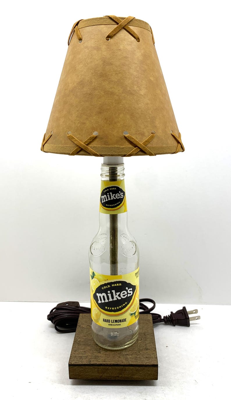May include: A unique table lamp made from a repurposed Mike's Hard Lemonade bottle. It features a tan lampshade with decorative stitching and a wooden base. The lamp is plugged into a standard US electrical outlet.