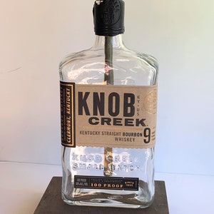 May include: A clear glass bottle lamp with a gold-colored lampshade. The bottle is labeled "Knob Creek Kentucky Straight Bourbon Whiskey" and "100 Proof". The lamp is sitting on a dark brown wooden base and has a yellow cord with a plug.