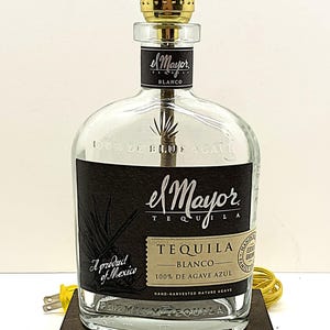 May include: A unique lamp made from an "el Mayor Tequila Blanco" bottle. The clear glass bottle features a black label with white text and a gold lamp fixture. The lamp sits on a dark brown wooden base. The lamp is plugged in with a yellow cord.