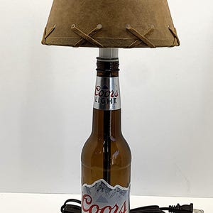 May include: A unique lamp crafted from a Coors Light beer bottle. The brown glass bottle base supports a tan lampshade with decorative stitching. The lamp sits on a small wooden base and has a black power cord.