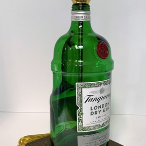 Op de afbeelding: Een groene glazen fles met een gouden lampenkap erbovenop. De fles is gelabeld met "Tanqueray London Dry Gin Est. 1830". De fles staat op een donkerbruine houten basis.