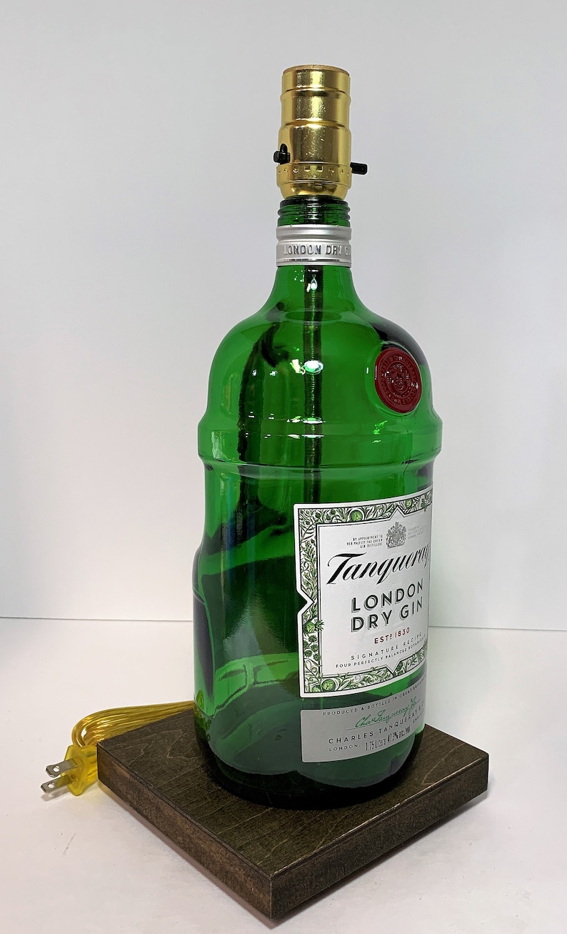Op de afbeelding: Een groene glazen fles met een gouden lampenkap erbovenop. De fles is gelabeld met "Tanqueray London Dry Gin Est. 1830". De fles staat op een donkerbruine houten basis.