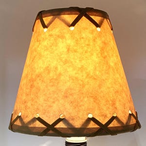 May include: A lampshade with a textured, tan-colored surface and a dark brown trim. The trim features an X-shaped design with small, evenly spaced holes. The lampshade is lit from within, creating a warm glow.