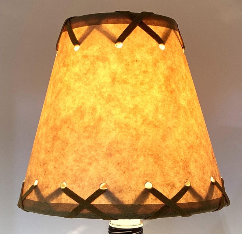 May include: A lampshade with a textured, tan-colored surface and a dark brown trim. The trim features an X-shaped design with small, evenly spaced holes. The lampshade is lit from within, creating a warm glow.