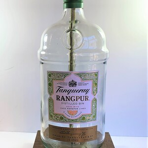 May include: A clear glass bottle lamp with a gold colored lampshade and a green base. The bottle is labeled "Tanqueray Rangpur. Distilled Gin. Made with Rare Rangpur Limes & Other Botanicals. Produced & Bottled in Great Britain. Charles Tanqueray & Co. London England."