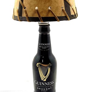 May include: A unique lamp made from a Guinness Draught Stout bottle. The lamp features a brown lampshade with dark brown trim and decorative stitching. The base is a wooden block, and the lamp has a standard electrical plug.