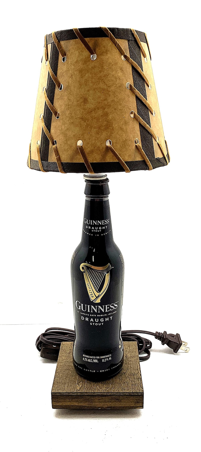 May include: A unique lamp made from a Guinness Draught Stout bottle. The lamp features a brown lampshade with dark brown trim and decorative stitching. The base is a wooden block, and the lamp has a standard electrical plug.