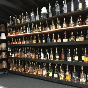 May include: A wall of shelves filled with glass bottles, each with a light bulb inside, creating a unique lighting display. The bottles are arranged in rows and columns, with a variety of shapes and sizes. The shelves are black and the bottles are clear glass.