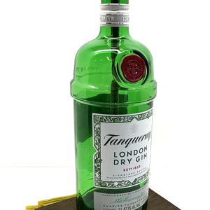 May include: A green glass bottle with a gold lampshade on top. The bottle is labeled "Tanqueray London Dry Gin" and has a red seal. The bottle is sitting on a wooden base.