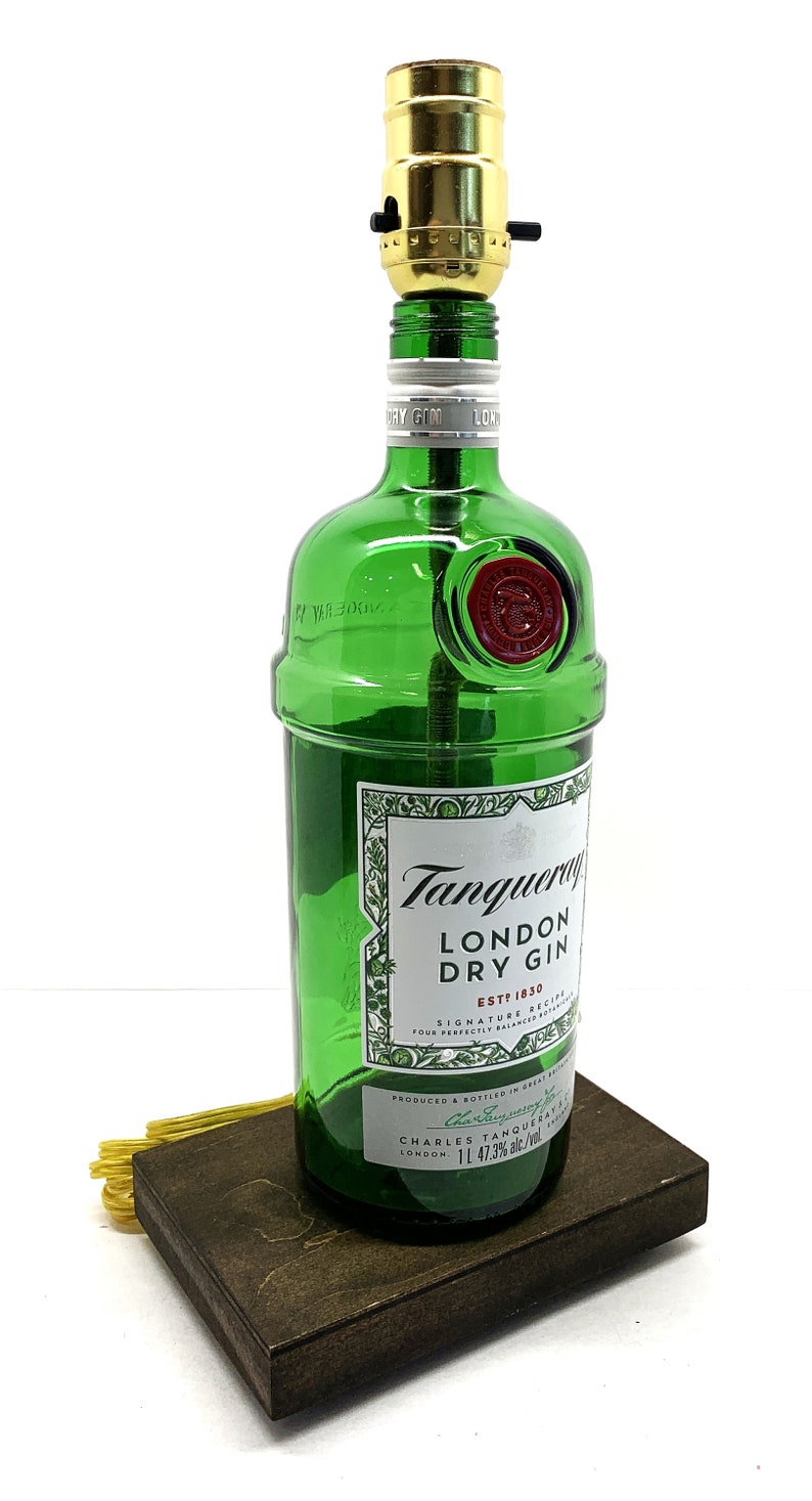 May include: A green glass bottle with a gold lampshade on top. The bottle is labeled "Tanqueray London Dry Gin" and has a red seal. The bottle is sitting on a wooden base.