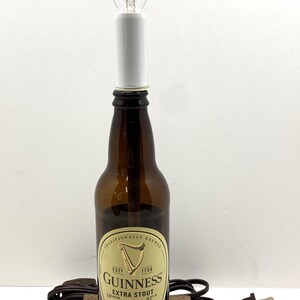 May include: A repurposed Guinness Extra Stout beer bottle lamp with a clear light bulb. The brown bottle sits on a wooden base, with a brown cord and plug. The label reads "Guinness Extra Stout".