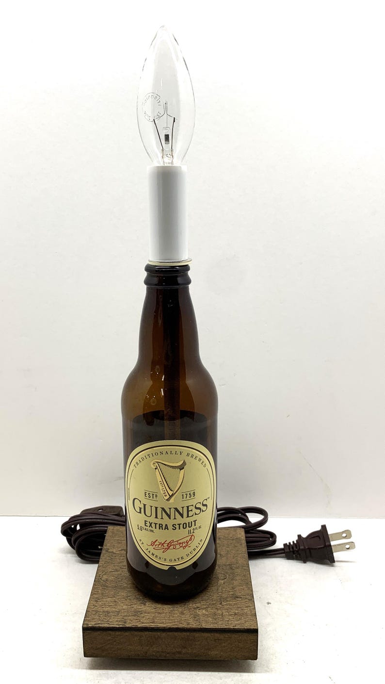 May include: A repurposed Guinness Extra Stout beer bottle lamp with a clear light bulb. The brown bottle sits on a wooden base, with a brown cord and plug. The label reads "Guinness Extra Stout".