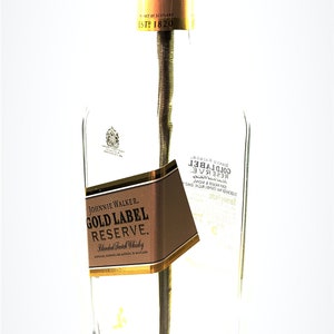 May include: A table lamp made from a repurposed Johnnie Walker Gold Label Reserve bottle. The bottle is clear glass with a brown label that reads "Johnnie Walker Gold Label Reserve Blended Scotch Whisky". The lamp has a gold metal socket and a brown wooden base.