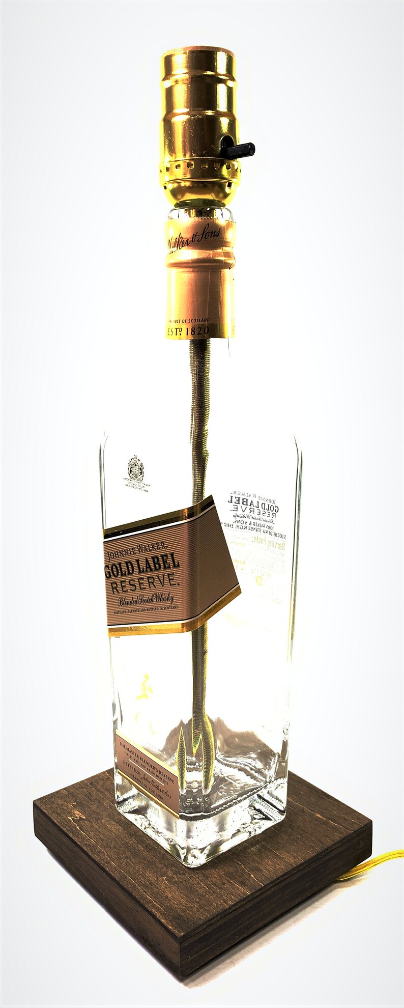 May include: A table lamp made from a repurposed Johnnie Walker Gold Label Reserve bottle. The bottle is clear glass with a brown label that reads "Johnnie Walker Gold Label Reserve Blended Scotch Whisky". The lamp has a gold metal socket and a brown wooden base.