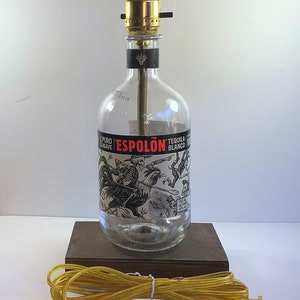 May include: A clear glass bottle lamp with a gold metal lampshade. The bottle has a black and white label with the text "El Puro Agave Espolón Tequila Blanco". The lamp is sitting on a wooden base and has a yellow cord with a plug.