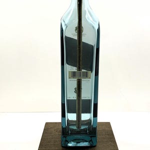 K&ouml;nnte beinhalten: Eine umfunktionierte Lampe aus einer blauen Spirituosenflasche, mit einem goldfarbenen Lampenhalter und einem braunen Holzsockel. Die Lampe hat ein gelbes Netzkabel. Die Flasche hat ein Etikett mit Text.