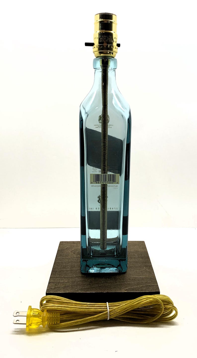 K&ouml;nnte beinhalten: Eine umfunktionierte Lampe aus einer blauen Spirituosenflasche, mit einem goldfarbenen Lampenhalter und einem braunen Holzsockel. Die Lampe hat ein gelbes Netzkabel. Die Flasche hat ein Etikett mit Text.