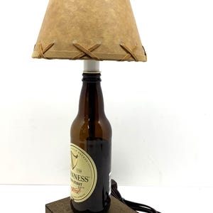May include: A handcrafted lamp made from a Guinness stout beer bottle. The lamp features a brown lampshade with decorative stitching and a wooden base. The lamp is plugged in and ready to use.