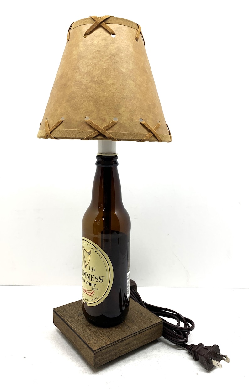May include: A handcrafted lamp made from a Guinness stout beer bottle. The lamp features a brown lampshade with decorative stitching and a wooden base. The lamp is plugged in and ready to use.