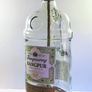 May include: A clear glass bottle lamp with a gold metal lampshade. The bottle has a label that reads "Tanqueray Rangpur Distilled Gin Made with Rare Rangpur Limes Other Botanicals Bottled in Great Britain Charles Tanqueray & Co England."
