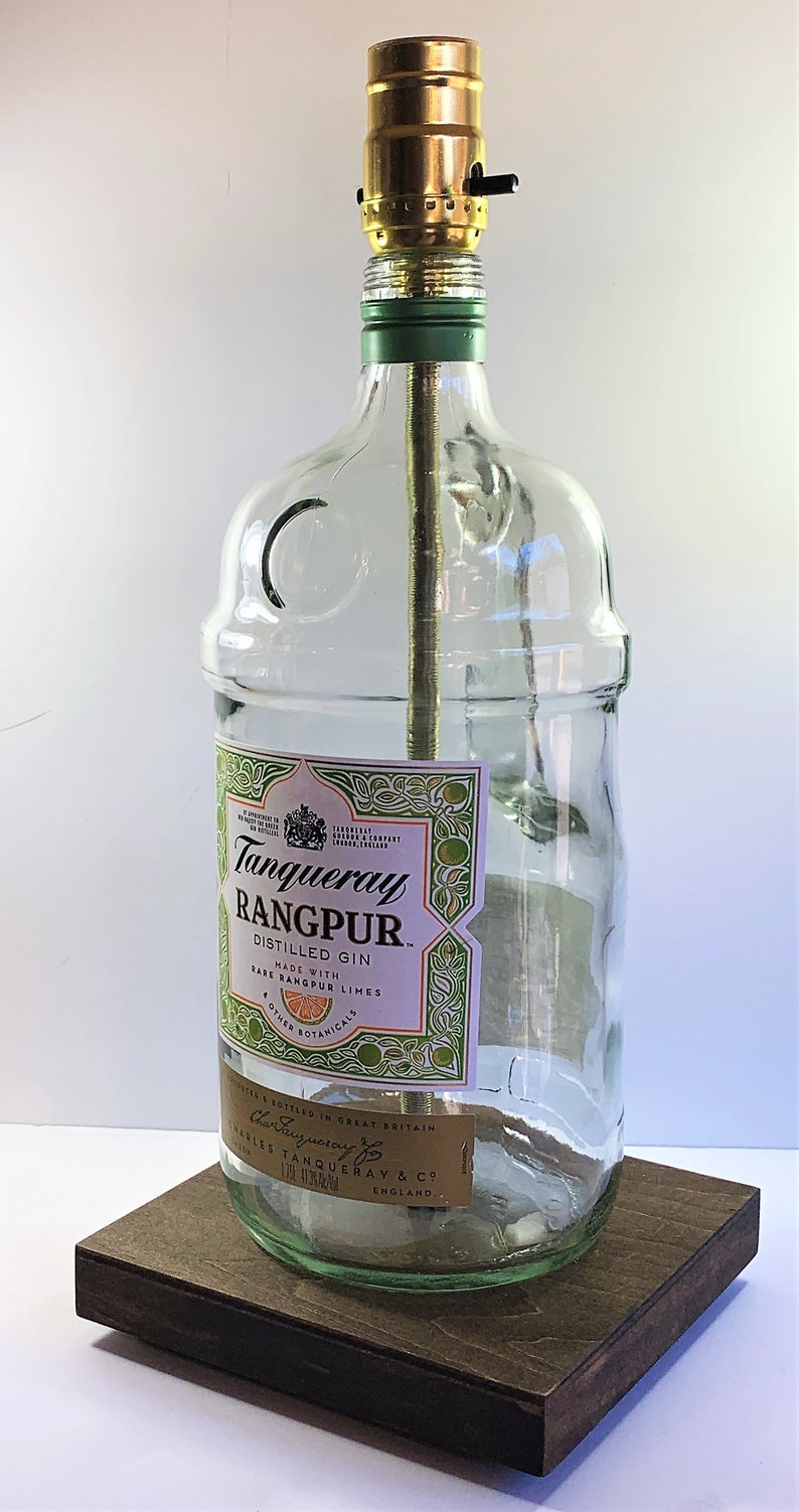 May include: A clear glass bottle lamp with a gold metal lampshade. The bottle has a label that reads "Tanqueray Rangpur Distilled Gin Made with Rare Rangpur Limes Other Botanicals Bottled in Great Britain Charles Tanqueray & Co England."