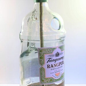 May include: A clear glass bottle lamp with a gold metal lampshade and a wooden base. The bottle is labeled with the text "Tanqueray Rangpur Distilled Gin Made with Rare Rangpur Limes & Other Botanicals Produced & Bottled in Great Britain Charles Tanqueray & Co. London 1.75L 41.3% alc./vol."