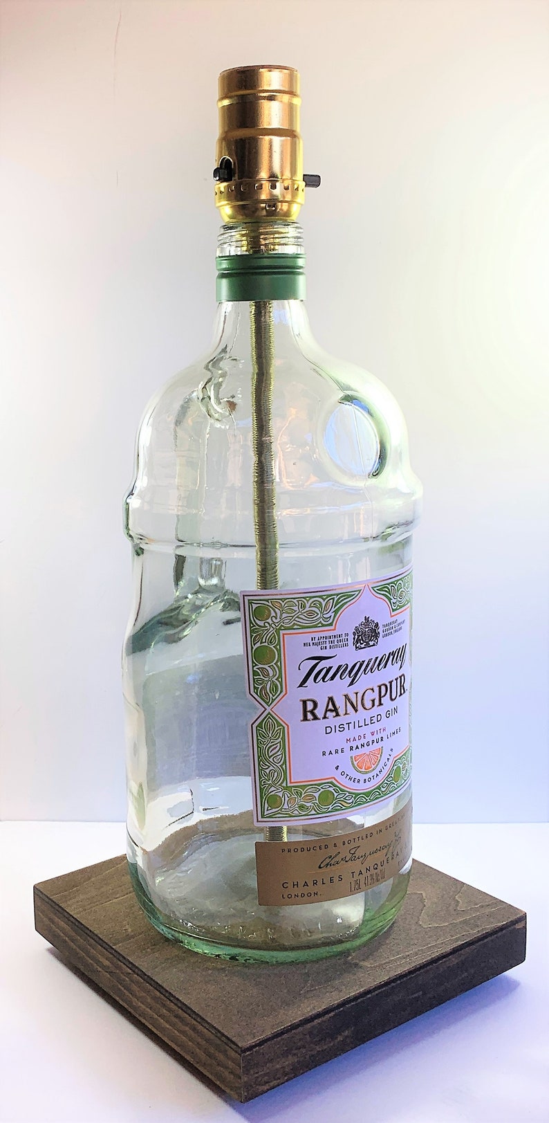 May include: A clear glass bottle lamp with a gold metal lampshade and a wooden base. The bottle is labeled with the text "Tanqueray Rangpur Distilled Gin Made with Rare Rangpur Limes & Other Botanicals Produced & Bottled in Great Britain Charles Tanqueray & Co. London 1.75L 41.3% alc./vol."