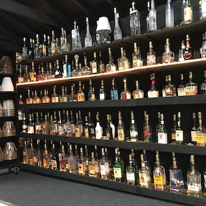 May include: A wall of shelves filled with glass bottles, each with a light bulb inside, creating a unique lighting display. The bottles are arranged in rows and columns, with a variety of shapes and sizes. The shelves are black and the bottles are clear glass.