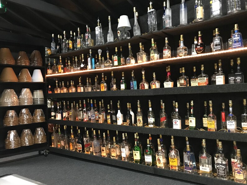 May include: A wall of shelves filled with glass bottles, each with a light bulb inside, creating a unique lighting display. The bottles are arranged in rows and columns, with a variety of shapes and sizes. The shelves are black and the bottles are clear glass.