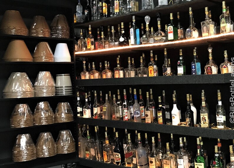 May include: A wall of shelves filled with glass bottles, each with a gold top. The bottles are arranged in rows and columns, and they are all different sizes and shapes. There are also several stacks of lampshades in the image, which are brown and white.