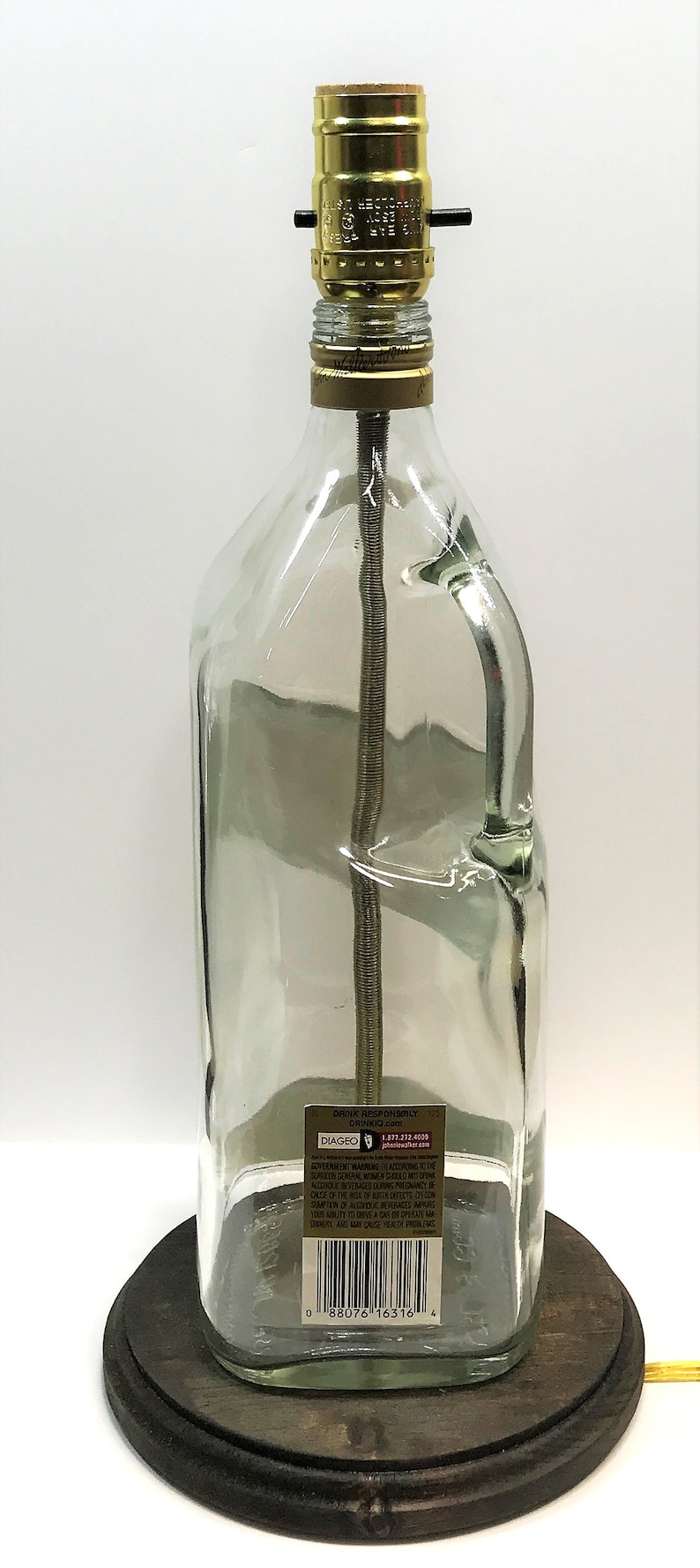 Puede incluir: Una l&aacute;mpara de botella de vidrio transparente con una pantalla de metal dorado y una base de madera. La botella tiene una etiqueta que dice "Drink Responsibly" y "Diageo".