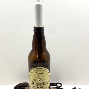 May include: A repurposed Guinness Extra Stout beer bottle lamp. The brown glass bottle features the Guinness label and is mounted on a wooden base. A white socket holds a clear, pointed light bulb. A brown electrical cord and plug are attached.