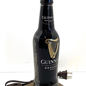 May include: A repurposed Guinness Draught Stout bottle lamp. The black bottle features the Guinness logo and text, with a white lamp shade. It sits on a square wooden base with a brown cord and plug.