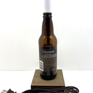 May include: A repurposed Guinness Extra Stout beer bottle lamp. The brown glass bottle has a white light fixture and sits on a small wooden base. A brown electrical cord with a plug is attached.