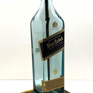 K&ouml;nnte beinhalten: Eine umfunktionierte Lampe aus einer Johnnie Walker Blue Label Whiskyflasche. Die blaue Glasflasche hat ein gold-schwarzes Etikett und steht auf einem dunklen Holzsockel. Die Lampe hat eine goldfarbene Fassung und ein gelbes Kabel.