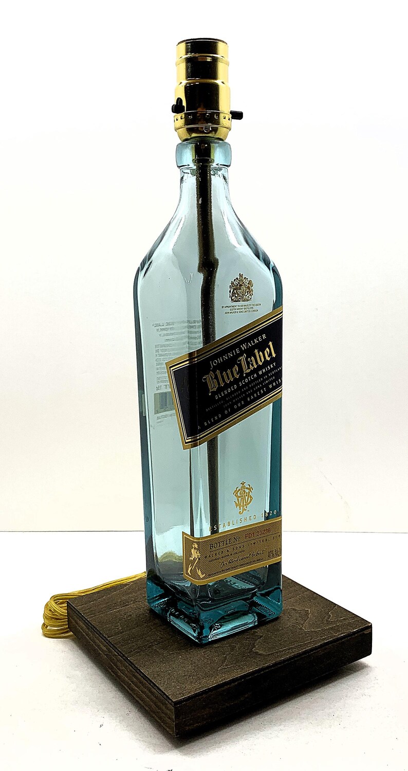 K&ouml;nnte beinhalten: Eine umfunktionierte Lampe aus einer Johnnie Walker Blue Label Whiskyflasche. Die blaue Glasflasche hat ein gold-schwarzes Etikett und steht auf einem dunklen Holzsockel. Die Lampe hat eine goldfarbene Fassung und ein gelbes Kabel.